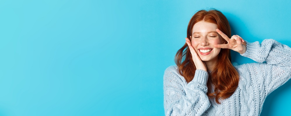 Devojka ridje kose se smeje i vidi se estetika lepih belih zuba i jednom rukom pridrzava lice drugom pokazuje slovo v kao pobeda 