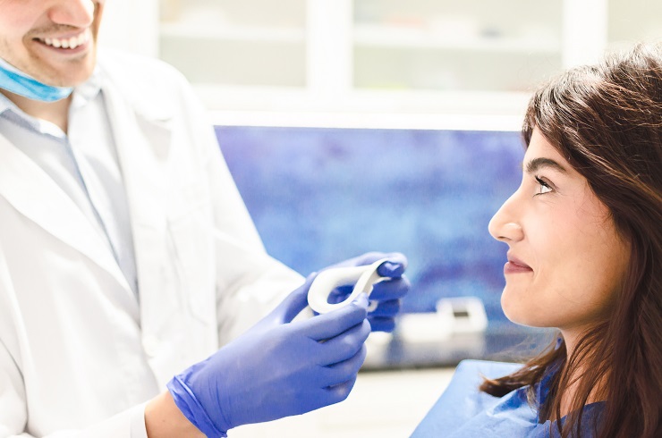 Devojka brineta sedi na zubarskoj stolici i ceka dok joj doktor prinosi ekarter za izbeljivanje zuba